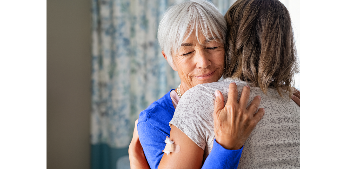 An older woman with white hair smiles while embracing another person; she has a bandage on her arm