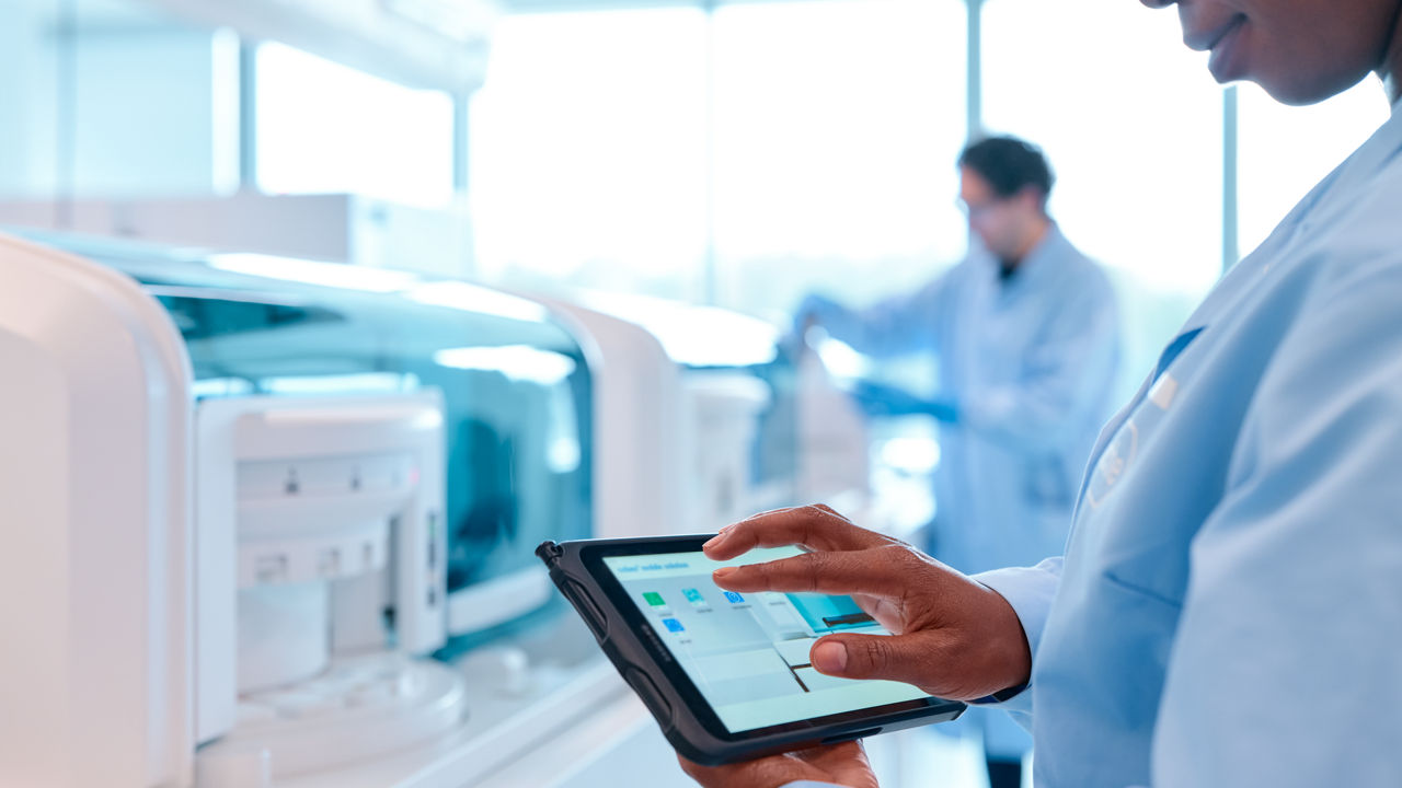 Technician in a lab coat in front of a dîagnsotics instrument holds a smart device showing digital solutions for core lab