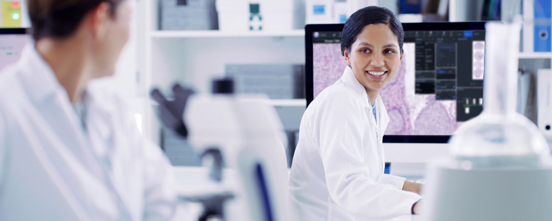 two lab pathologists exchange smiles - the pathologist in focus is reviewing images of stained tissue sections on her computer