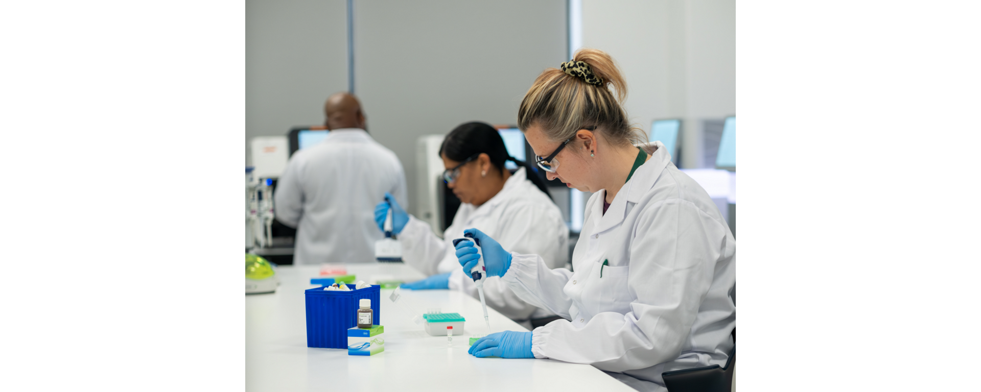 Two technicians wearing lab coats sitting at a laboratory bench pipetting adapters or beads during NGS library preparation