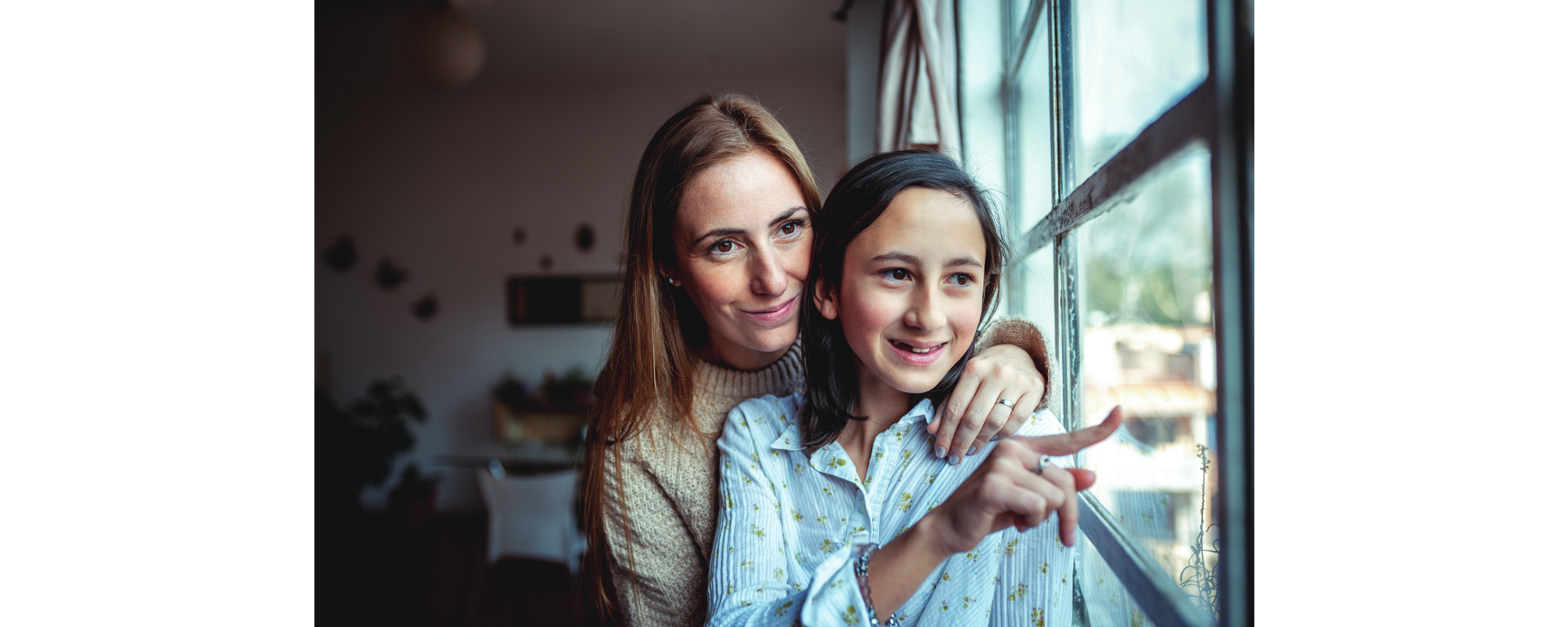 A mother and her young daughter stand side by side gazing out of a large window