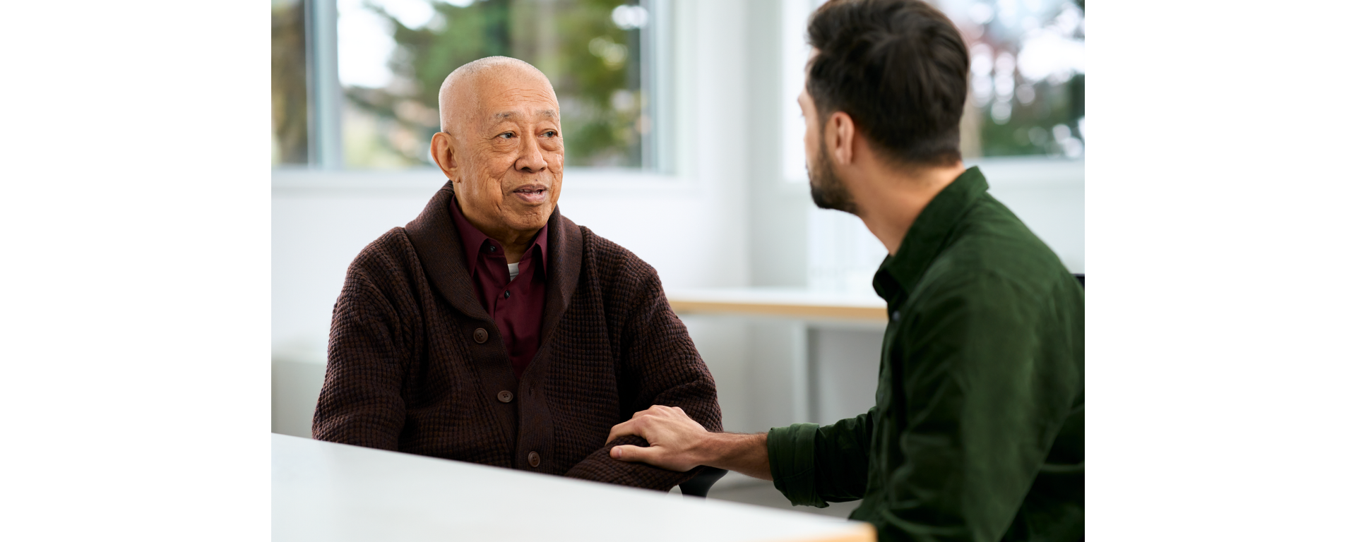 A young man in a green shirt places a comforting hand on the arm of an older man in a brown cardigan