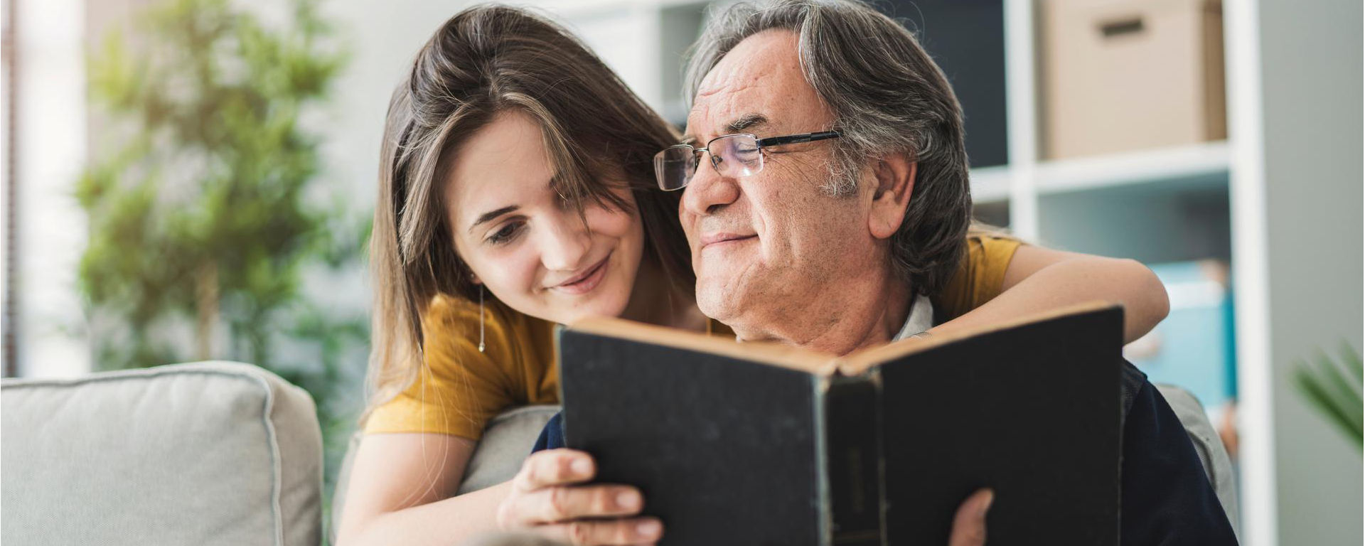 Father sits reading his book and his daughter hugs him from behind the couch They are both smiling