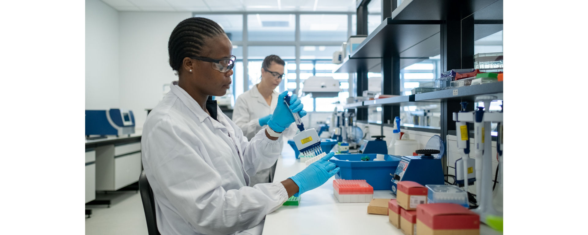A scientist in a lab coat and blue gloves uses a pipette at a workbench
